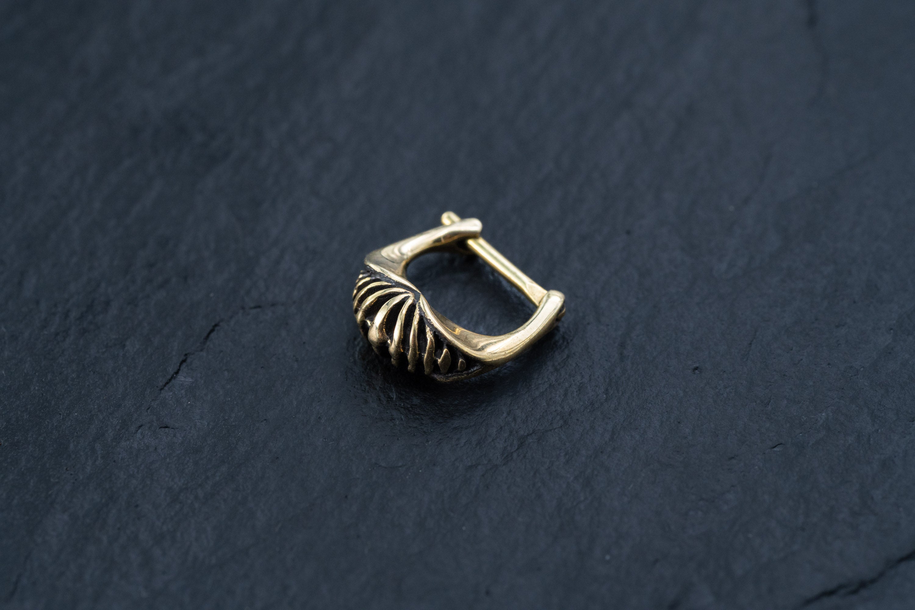 A close-up photograph of the Kaya Movement DIARA Biomechanical Clicker Hoop Septum Nose Ring in Gold, featuring a unique ribbed design and polished finish, placed on a dark, textured surface. The 16 gauge earring boasts an elegant hinged clasp, reminiscent of an exquisite cyborg clicker.